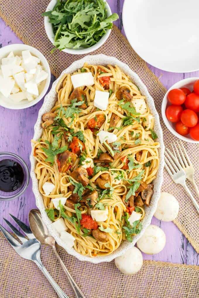 Roasted Mushroom Caprese Pasta in bowl with forks, spoon, bowl of tomatoes,, cheese, herbs and sauce on purple table with mushrooms