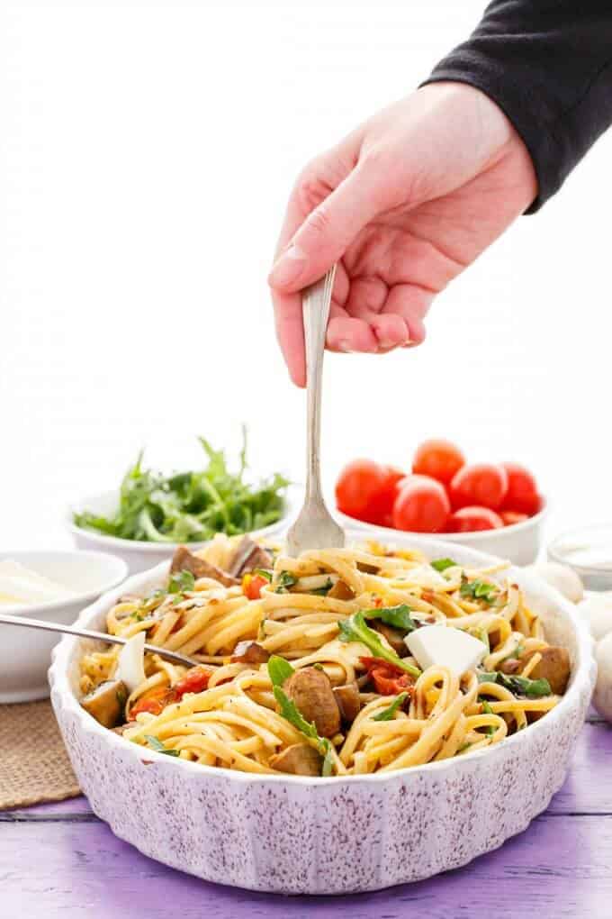 Roasted Mushroom Caprese Pasta in bowl with fork held by hand. Bowls of herbs, cheese and tomatoes in the background