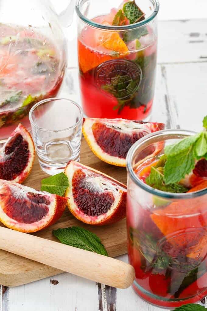 Blood Orange Mojitos in glass cups with bloodoranges, herbs, glass shot, wooden pad and tool and glass pitcher on the table