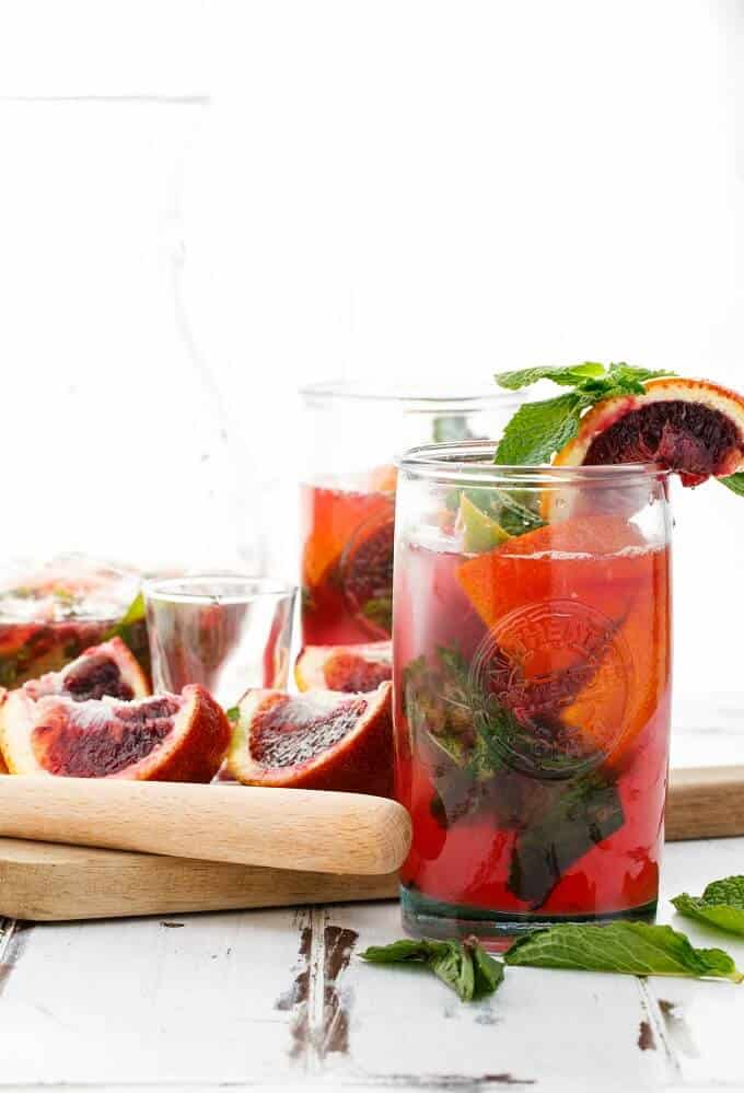 Blood Orange Mojitos in glass cups, glass pitcher with herbs, bloodoranges, wooden pad and wooden kitchen tool on the table