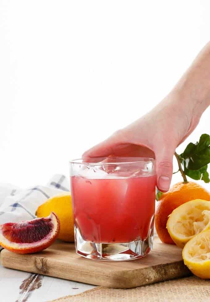 Blood Orange Whiskey Sour in glass cup held by hand on wooden pad with lemons, oranges and cloth wipe