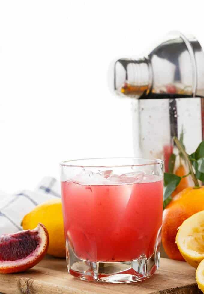 Blood Orange Whiskey Sour in glass cup with ice on wooden pad with lemons, oranges and cloth wipe. Cocktail shaker in the background