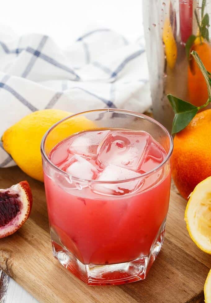 Blood Orange Whiskey Sour in glass cup with ice on wooden pad with oranges, lemons and cloth wipe