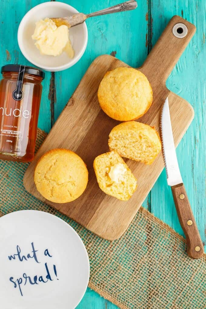 Buttermilk Honey Cornbread Muffins on wooden pad with knife. Jar of honey, bowl of butter with knife, white plate on blue table.