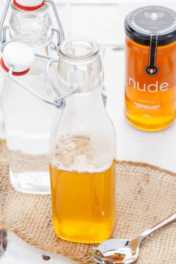 Homemade Honey Simple Syrup in glass bottle with empty glass bottle, spoon and jar of honey on white table