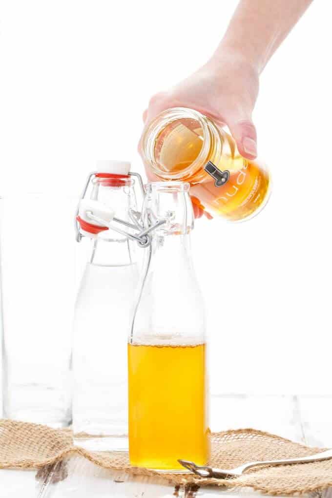 Homemade Honey Simple Syrup in glass bottle. Empty glass bottle, spoon, jar of honey held by hand over bottle of syrup on white table