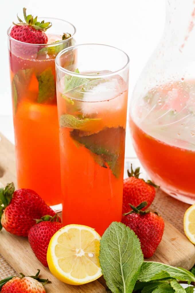 Homemade Strawberry Lemonade in glass cups and in glass pitcher on wooden pad with lemon, ripe strawberries and herbs