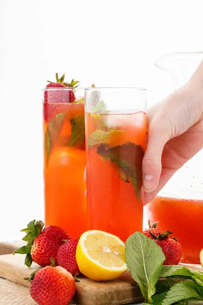 Homemade Strawberry Lemonade in glass cups held by hand .Lemon, ripe strawberries , herbs on wooden pad. Glass pitcher in the background