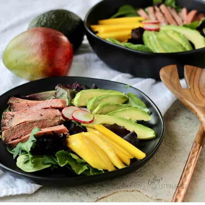 mango beef salad on black plate and in black bowl with wooden spatula, mango and avocado on the table