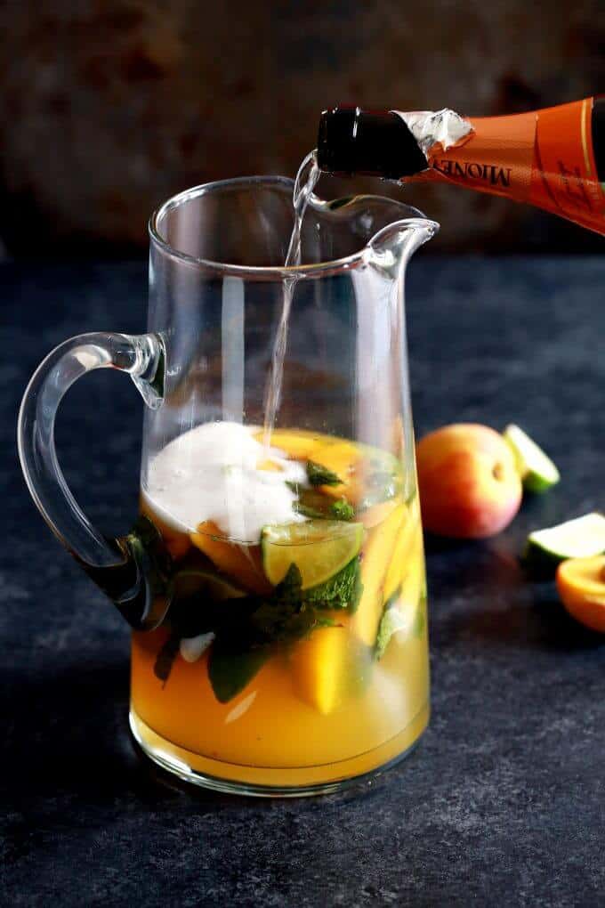 Peach prosseco mojito in glass pitcher, prosecco poured from bottle into pitcher, peaches and limes on the table