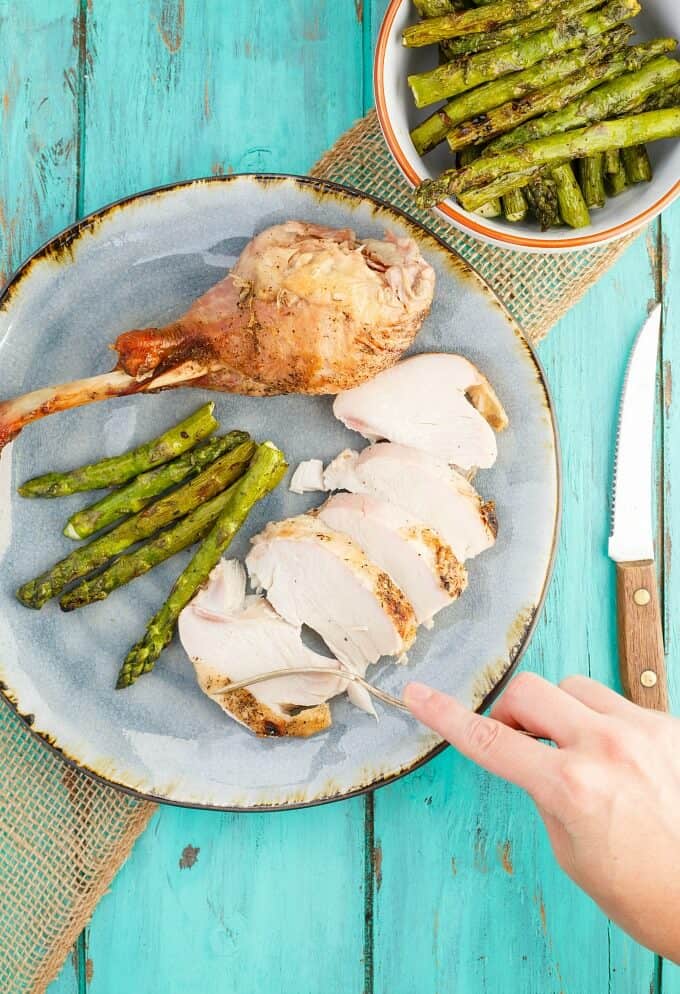 Grilled Spatchcock Turkey on blue plate with asparagus and fork held by hand. Asparagus in white orange bowl and knife on blue table