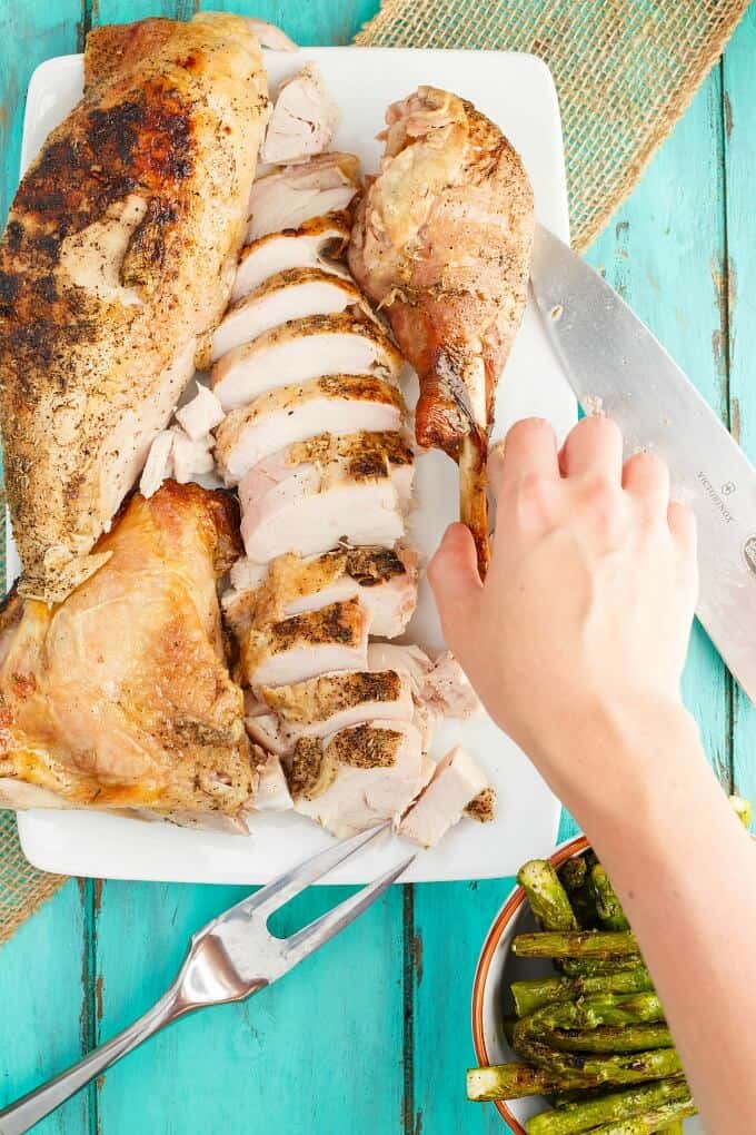 Grilled Spatchcock Turkey on white tray touched by hand. Knife, fork and asparagus in bowl on blue table