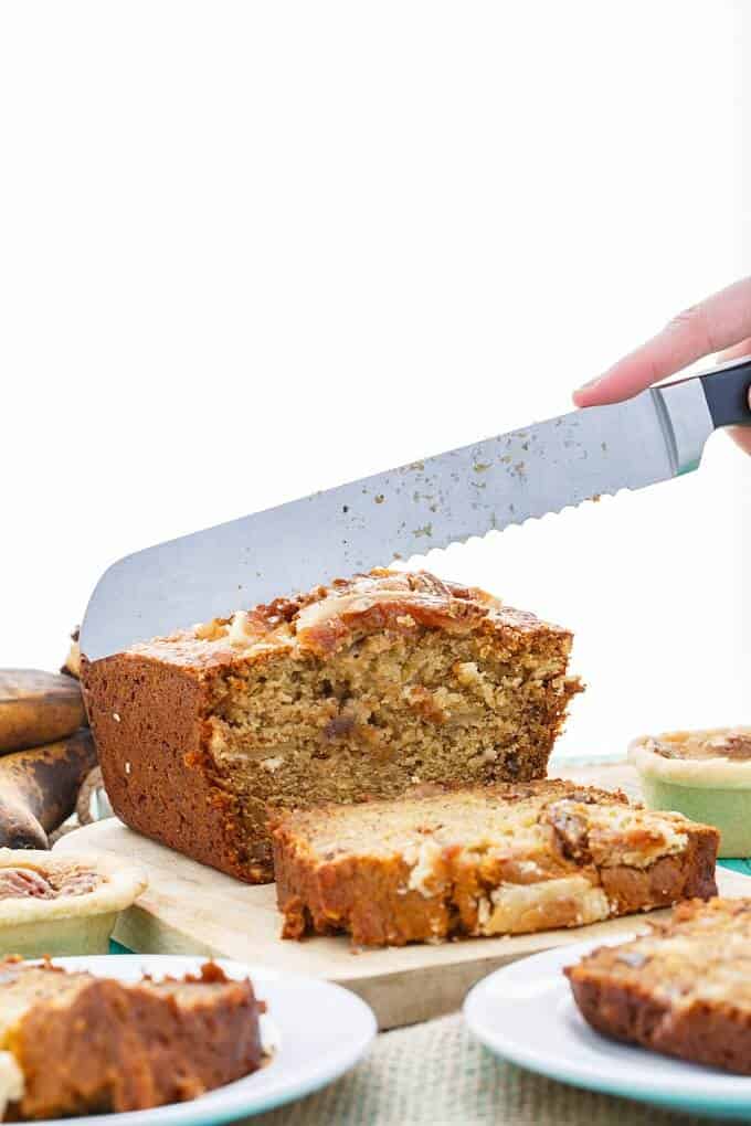 Homemade Butter Tart Banana Bread on wooden pad being sliced by knife held by hand. Plates with bread slices and small pies on table