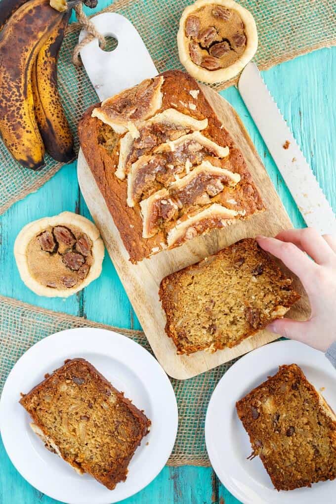 Homemade Butter Tart Banana Bread on wooden pad touched by hand and on white plates with slices of bread. Bananas, knife and small pies on blue table