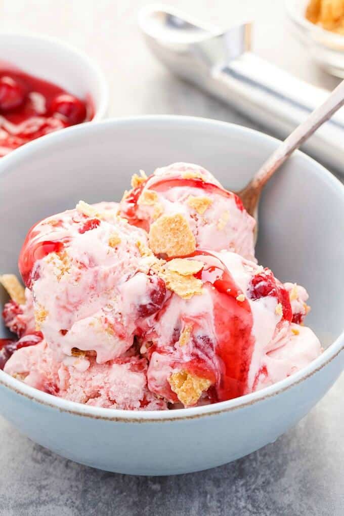Cherry Pie Ice Cream  in blue bowl with spoon. Ice cream spoon and bowl of sauce in the background
