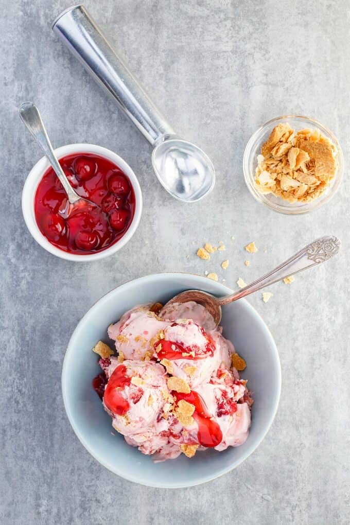 Cherry Pie Ice Cream  in blue bowl with spoon. Bowl of sauce with spoon and bowl of piece of pie and ice cream spoon on gray table