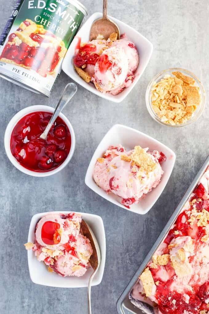 Cherry Pie Ice Cream  in black container and in white bowls with spoons. Bowl of sauce with spoon, bowl of cookies, cherries in can on gray table