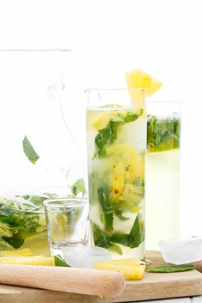 Fresh Pineapple Mojitos in glass cup on wooden pad with glass shot, ice cubes, herbs, pineapple slices, wooden tool. Glass pitcher in the background