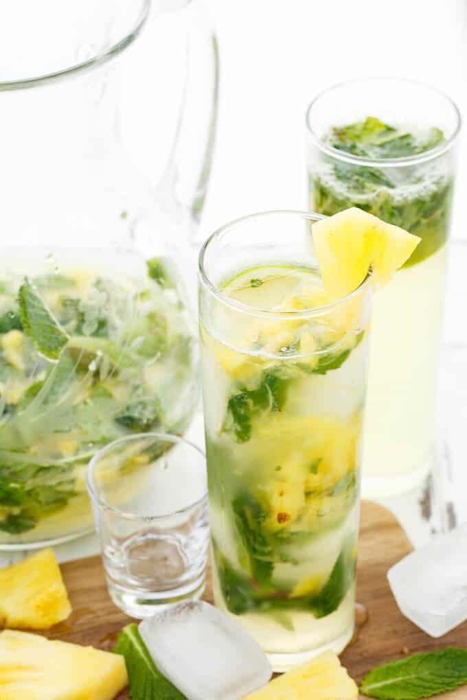 Fresh Pineapple Mojitos in glass cups and glass pitcher. Glass shot, ice cubes, herbs and pineapple  slices on wooden pad
