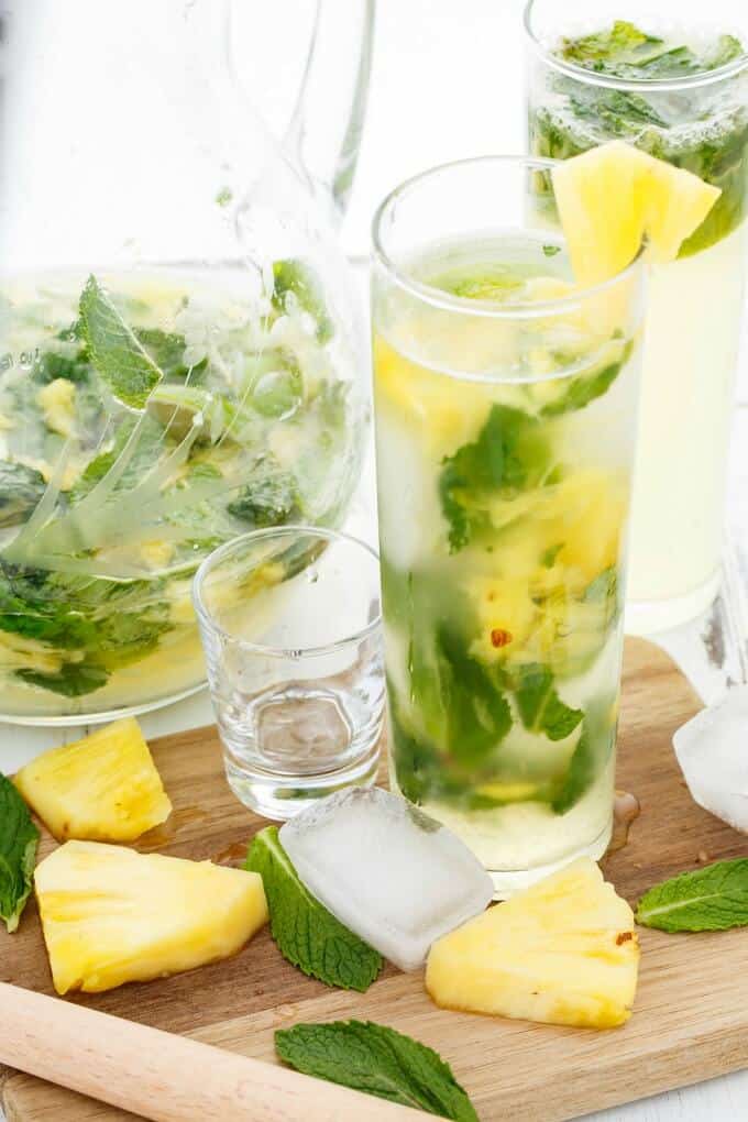 Fresh Pineapple Mojitos in glass cups, glass shot, ice cube, pineapple slices and herbs on wooden pad. Glass pitcher in the background