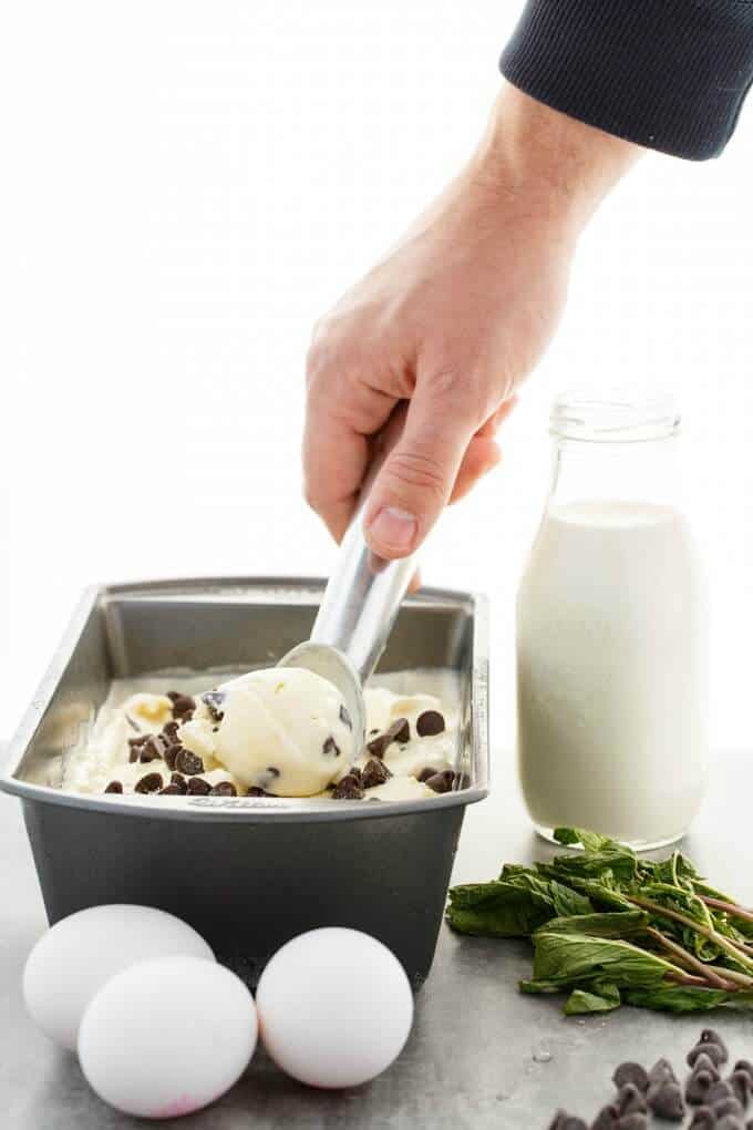 Homemade Fresh Mint Chocolate Chip Ice Cream in black cotainer with spoon held by hand. Boiled eggs, chocolate chips, herbs and glass jar of milk on the table