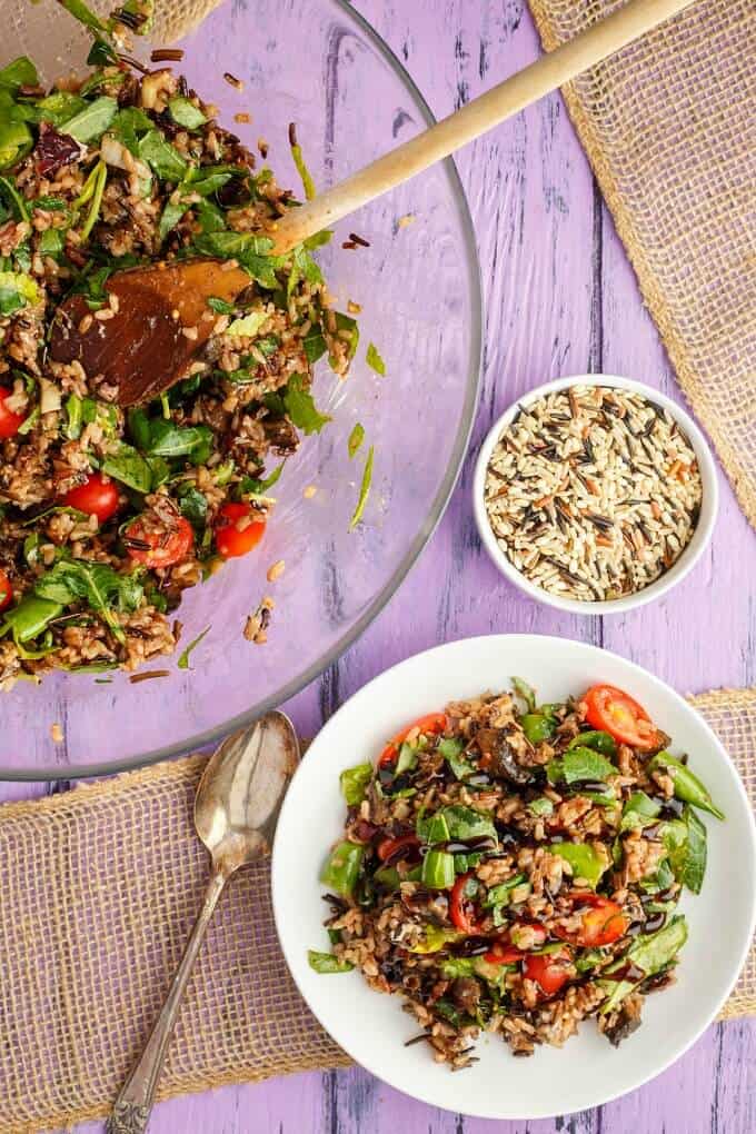 Roasted Mushroom Wild Rice Salad on white plate and in glass bowl with wooden spatula. Spoon and bowl of wild rice on purple table