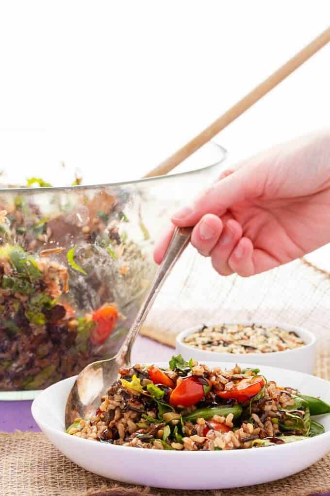 Roasted Mushroom Wild Rice Salad on white plate with spoon held by hand. Bowl of rice, glass bowl with salad and wooden spatula on table