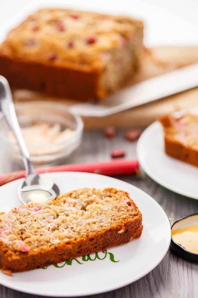 Honey-Rhubarb Banana Bread slices on white plates with spoon. Rhubarb, glass bowl, wooden pad with loaf of bread with knife on the table.