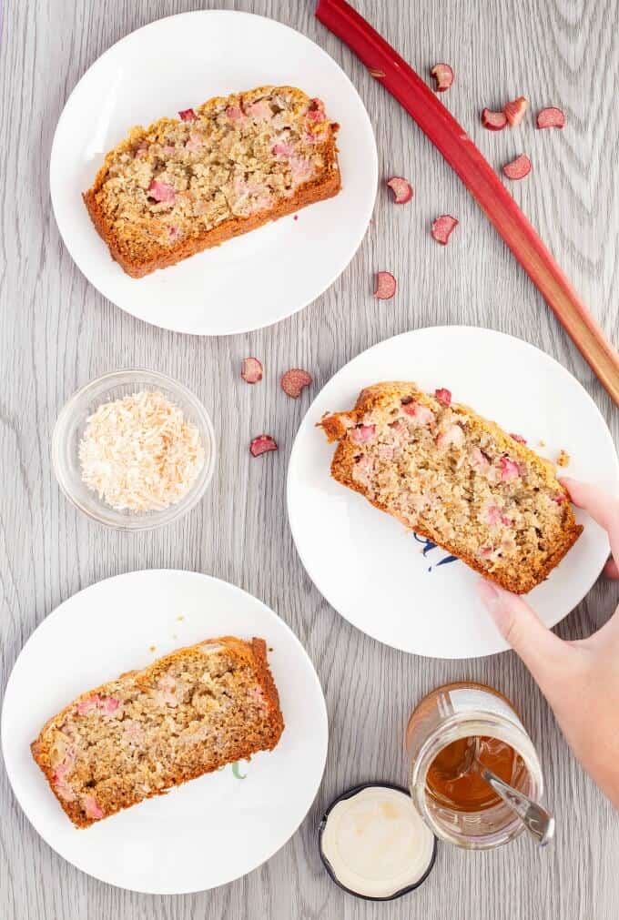 Honey rhubarb banana bread slices on white plates touched by hand. Rhubarb, honey jar with spoon, bowl of ingredient on gray table