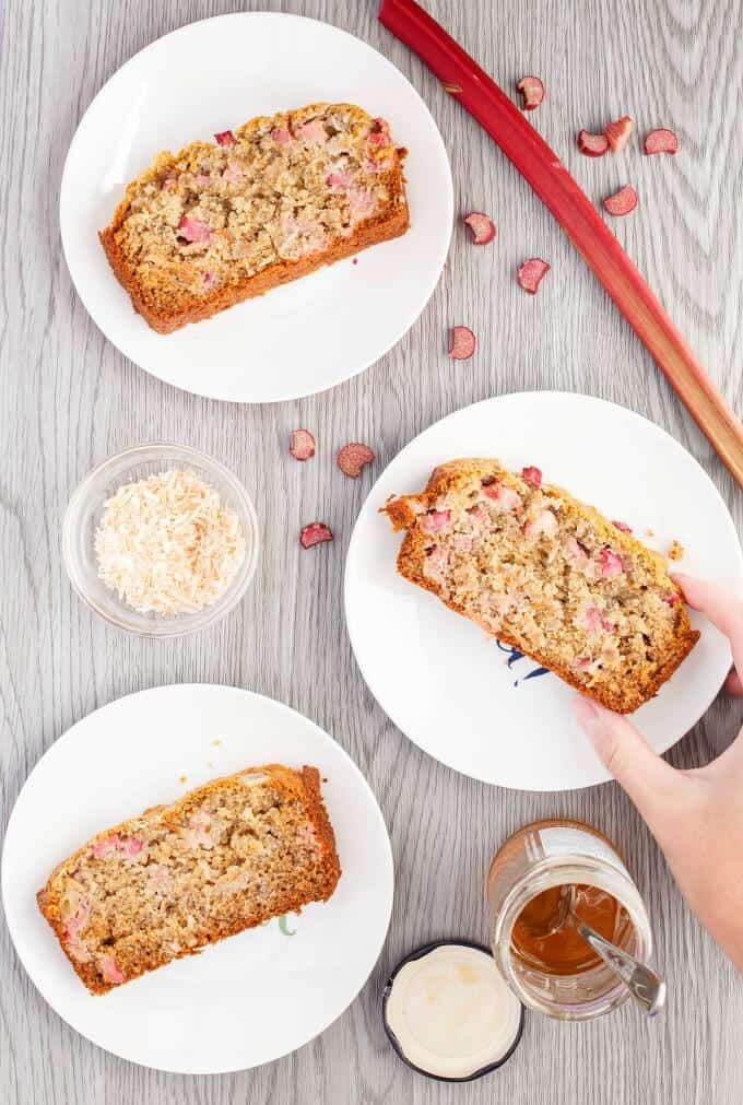 Honey-Rhubarb Banana Bread on white plates. One slice touched by hand. Rhubarb, honey jar and small glass bowl on brown table.