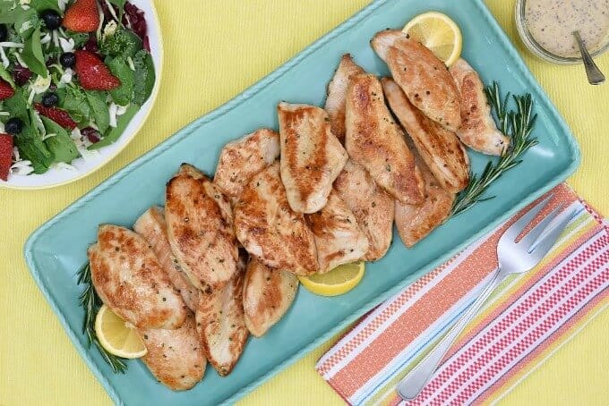 Honey lemon rosemary turkey tenders with herbs and lemons slices on blue tray. Fork, bowl with sauce, plate with veggies and fruits on cloth wipe on yellow table