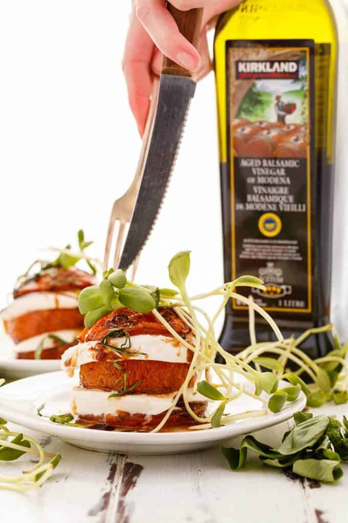 Tomato-Mozzarella Stacks on white plates with herbs. Balsamic vingear in glass bottle with herbs on the table. Hands holding knife and fork