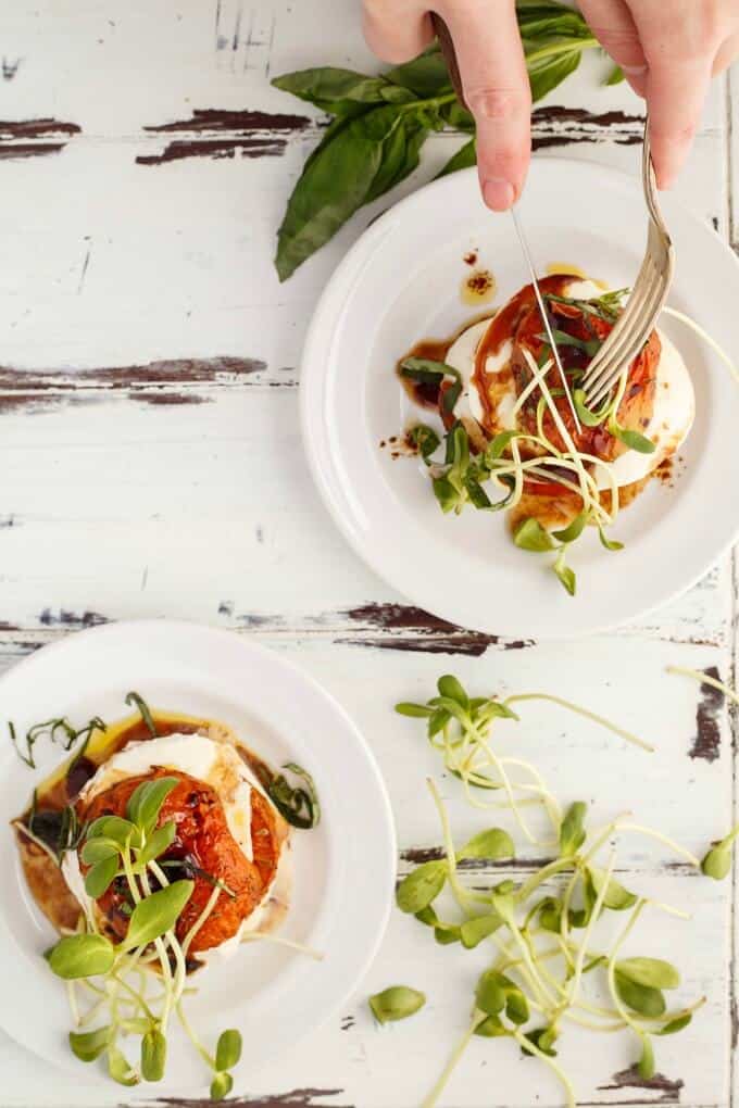 Tomato-Mozzarella Stacks  on white plates.with herbs on the table. Hands holding knife and fork over stack