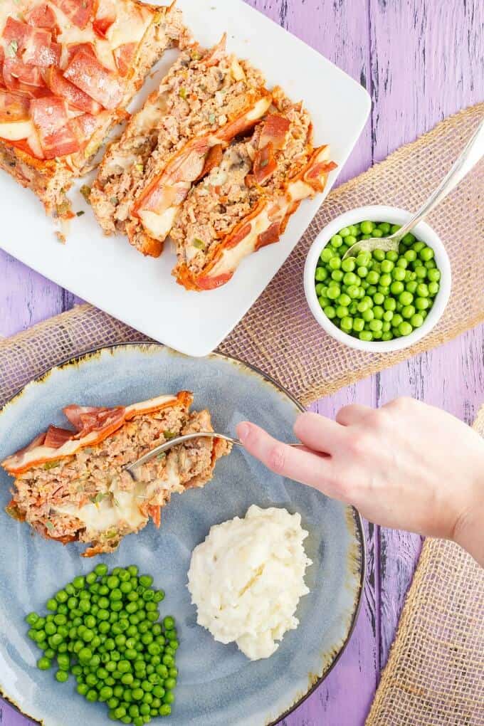 Turkey Pizza Meatloaf on blue plate with mashed potatoes, peas and fork held by hand. Turkey Pizza Meatloaf on white tray next to bowl of peas with spoon on purple table