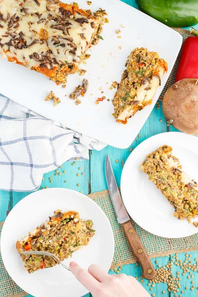 Vegetarian Pizza Meatloaf on white tray and white plates with fork held by hand. Knife, cloth wipe, mushrom and vegetable with scattered lentils on blue table