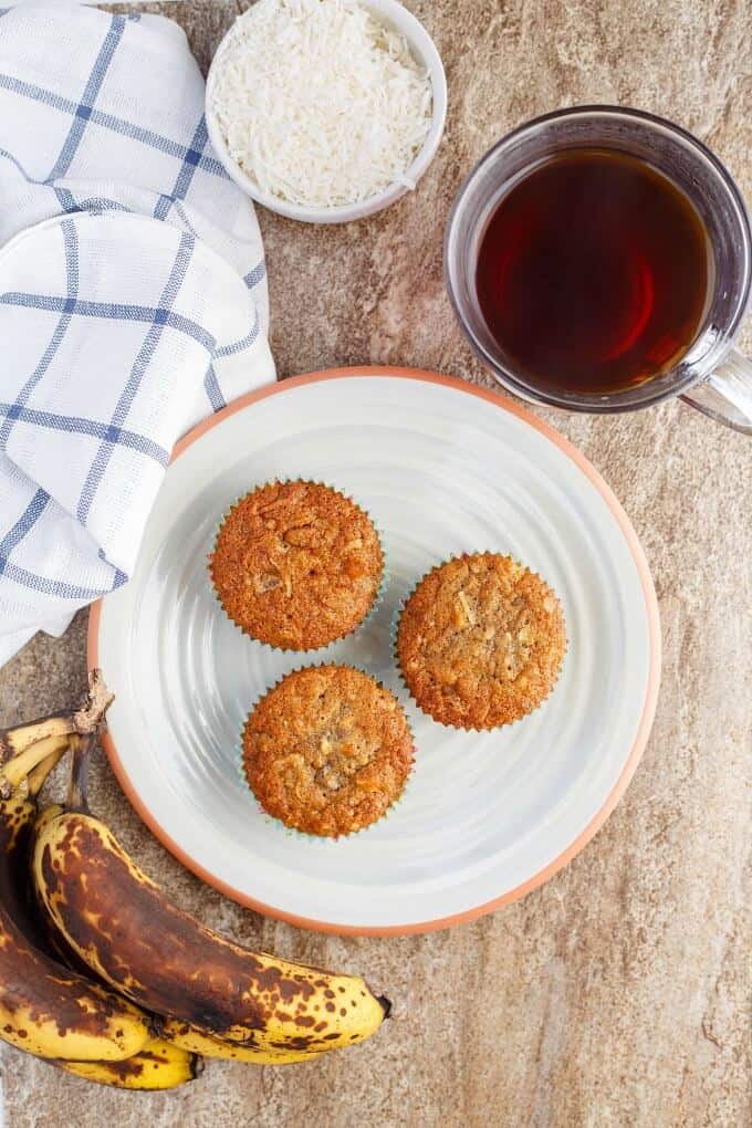 Chocolate Chip Banana Bread Muffins with Coconut & Nuts on white plate with bananas, bowl of sliced coconut, jar of juice and cloth wipe on brown table