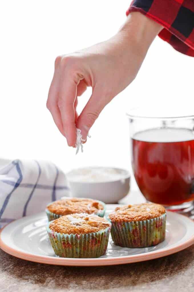 Chocolate Chip Banana Bread Muffins with Coconut & Nuts on plate being sprinkled  by coconut  by hand. Jar of juice, white bowl and cloth wipe in the background