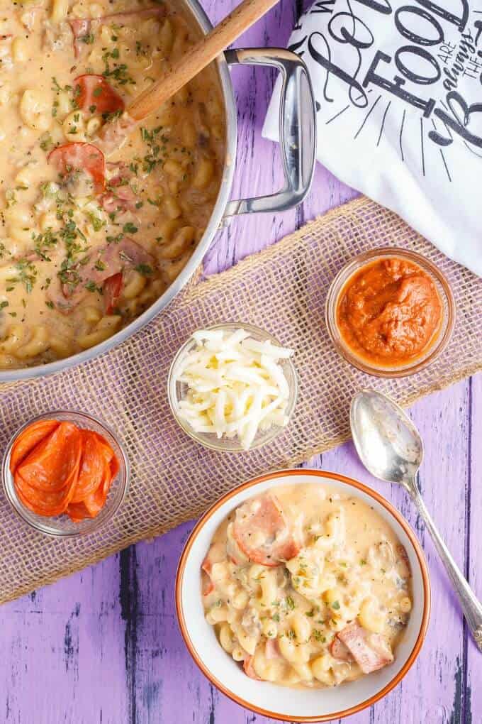 Homemade Pizza Mac and Cheese in white orange bowl and in cooking pot. Spoon, bowls with sauce, cheese and peperoni on purple table