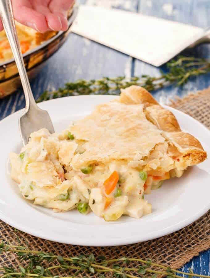 Homemade Turkey Pot Pie on white plate with fork held by hand. Herbs, spatula and glass tray with pie in the background