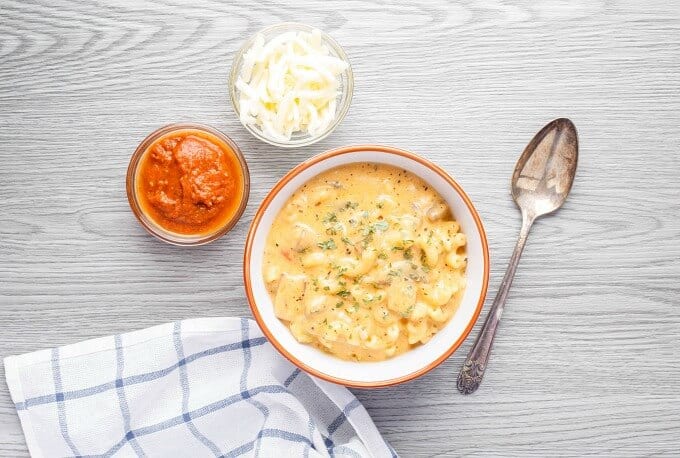 Homemade Vegetarian Pizza Mac and Cheese in white orange bowl. Spoon, bowls of sauce and cheese and cloth wipe on gray table