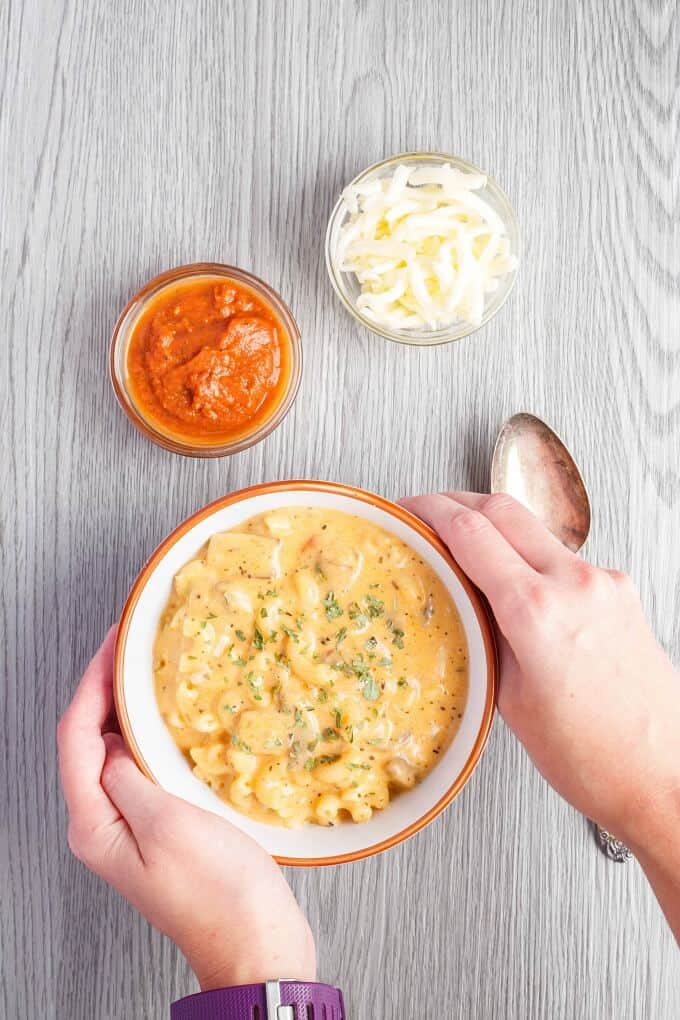 Homemade Vegetarian Pizza Mac and Cheese in white orange bowl held by hand, bowls of cheese and sauce with spoon on gray table