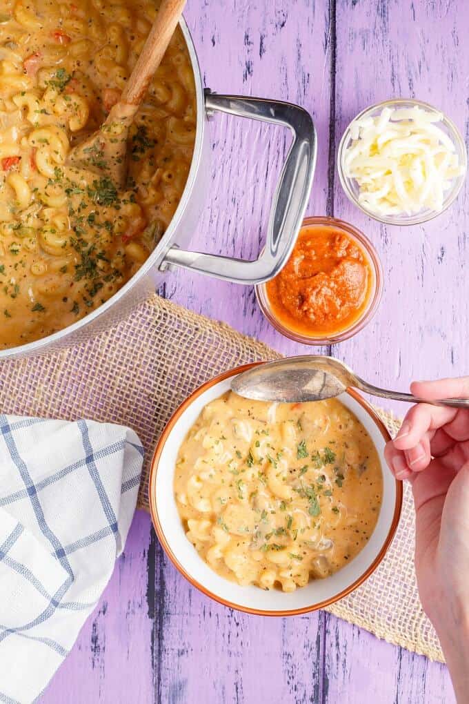 Homemade Vegetarian Pizza Mac and Cheese in white orange bowl with spoon held by hand, in pot with wooden spatula. Bowls with sauce and cheese , cloth wipe on purple table