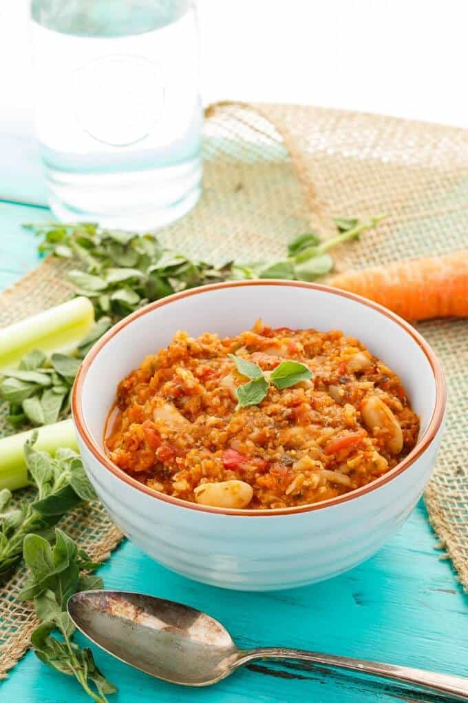 "Meaty" Vegetarian Chili  in white bowl with vegetable, spoon, herbs and glass cup on blue table