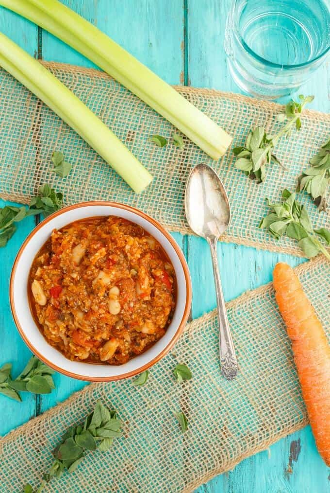 "Meaty" Vegetarian Chili  in white orange bowl with spoon, vegetable, herbs, and glass cup on blue table