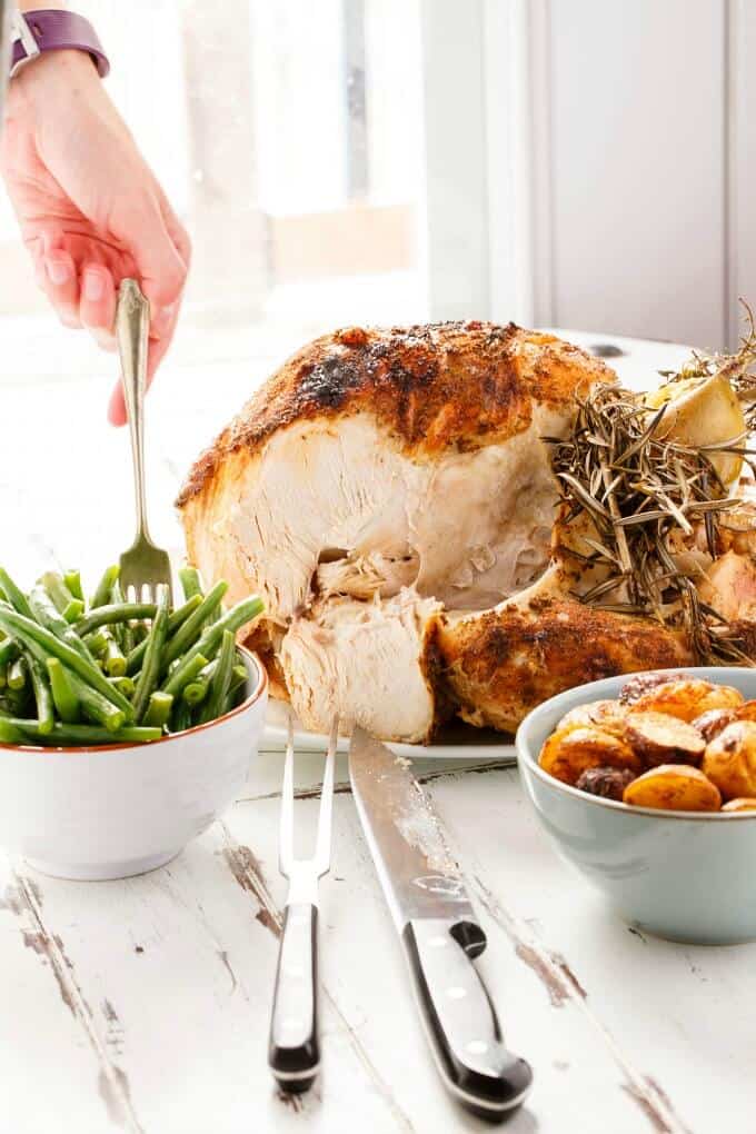 Slow Cooker Whole Turkey on white tray  with bowls of asparagus and potatoes. Fork held by hand over bowl, knife and fork on white table