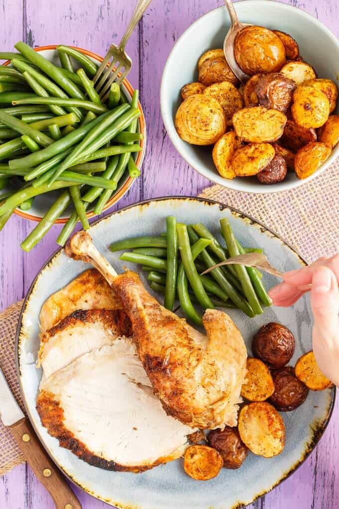 Slow Cooker Turkey on blue plate with asparagus and potaotes with fork over held by hand, bowl of asparagus with fork and bowl of potatoes with spoon, knife on purple table