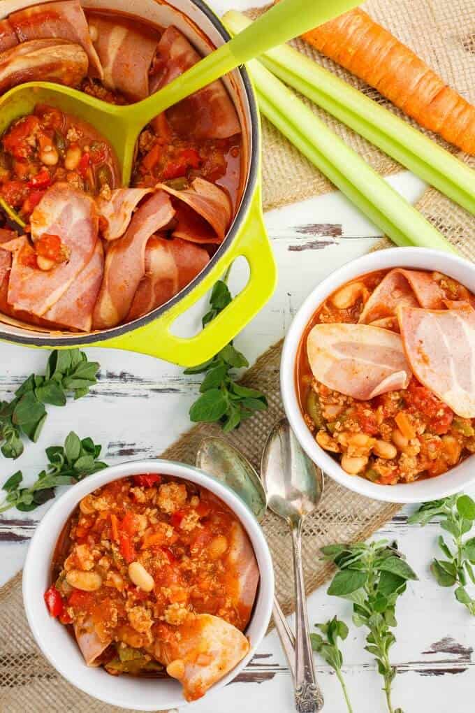 Turkey bacon chilli in white bowl and in green pot with herbs, spoons and vegetable on the table