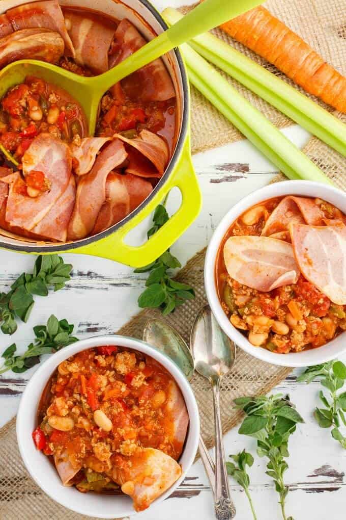 Turkey Bacon Chili in bowls and pot with green spatula. Vegetable, herbs and spoons on table