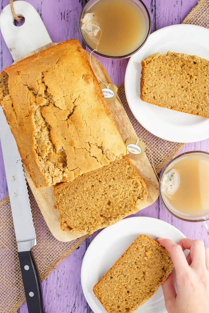 Earl Grey Tea Bread on wooden pad and white plates, one slice touched by hand. Cups of tea and knife on purple table