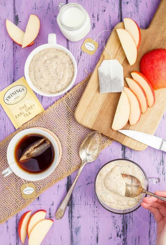 Homemade Earl Grey Pudding in cup with spoon held by hand. Wooden pad with apple slices and tea bag, Cup of tea,  tea bag, jar of milk, spoon, knife on purple table